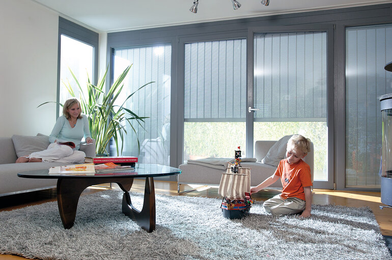 Wohnzimmer, mit großflächiger Fensterfront, das durch heruntergelassene s-onro Rollläden vor neugierigen Blicken und Sonneneinstrahlung geschützt ist.
