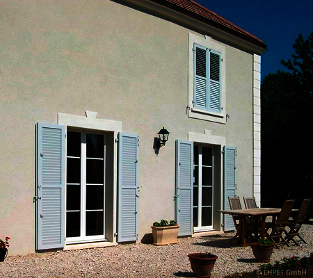 Schutz und Akzent: Haus mit bodentiefen Fenstern mit klassischen weißen Klappläden, beim Fenster im 1. O.G. geschlossen.