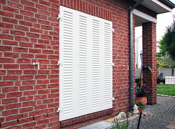 Geschlossene Klappläden in weiß vor einem bodentiefen Fenster eines roten Backsteinhauses