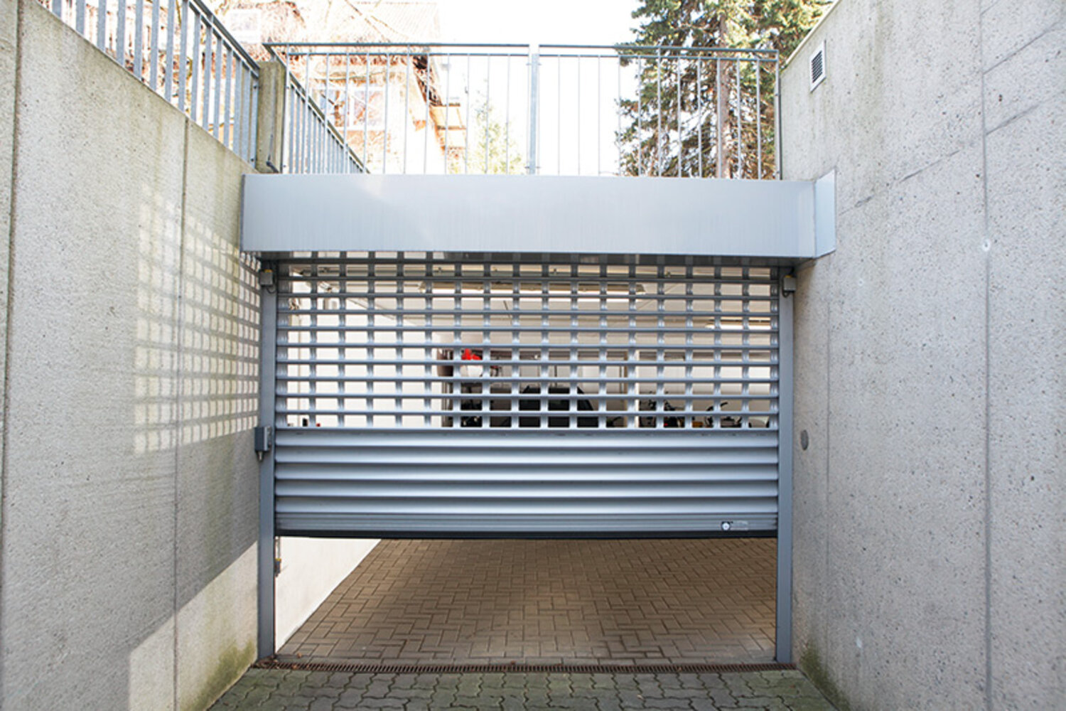 Ansicht eines halb heruntergefahrenen, alufarbenden Rolltores an einer Garageneinfahrt mit seitlichen Sichtbetonwänden Ansicht eines halb heruntergefahrenen, alufarbenden Rolltores an einer Garageneinfahrt mit seitlichen Sichtbetonwänden