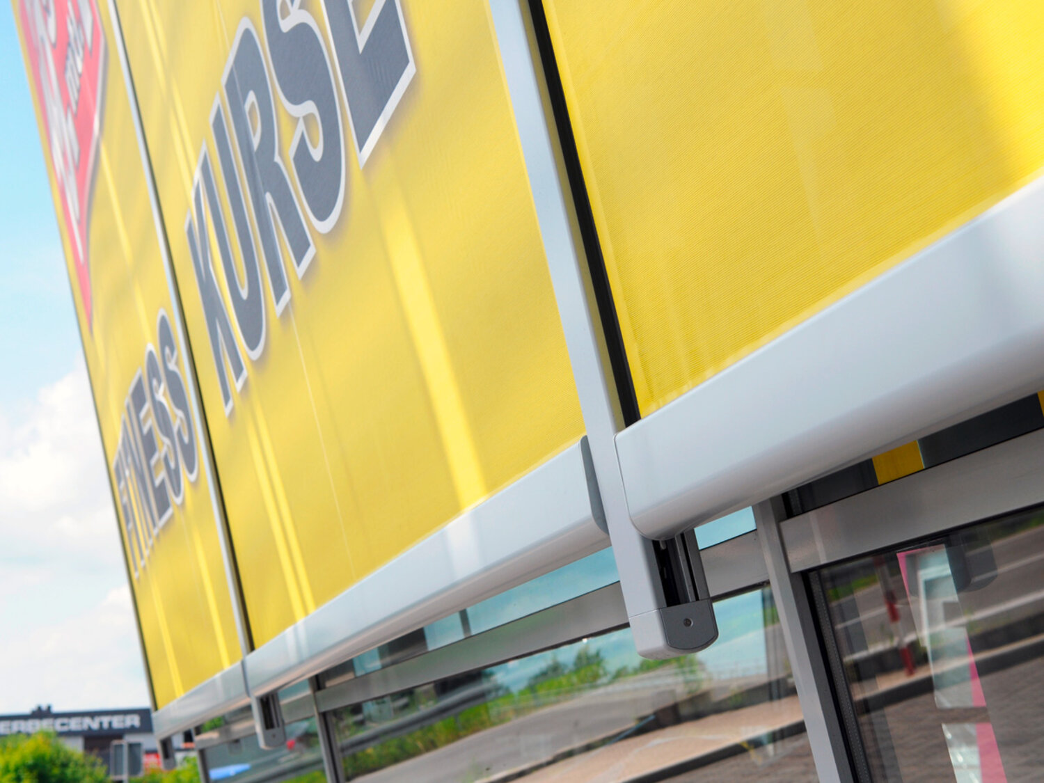 Detailansicht einer ausgefahrenen, gelben Fassadenmarkise, mit grauer Metallkonstruktion, die mit Werbung bedruckt ist Detailansicht einer ausgefahrenen, gelben Fassadenmarkise, mit grauer Metallkonstruktion, die mit Werbung bedruckt ist