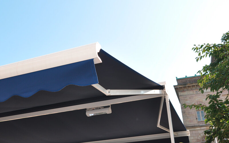 Detailansicht einer ausgefahrenen Doppelmarkise mit dunkelblauem Stoff und weißer Metallkonstruktion Detailansicht einer ausgefahrenen Doppelmarkise mit dunkelblauem Stoff und weißer Metallkonstruktion