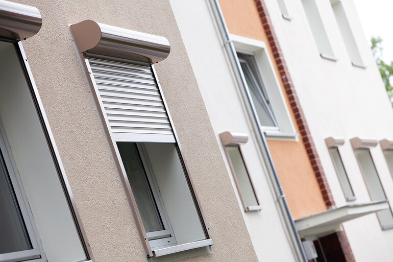 Nachträglich montierter, halb heruntergelassener aluminiumfarbender Vorbaurollladen am Fenster einer Wohnanlage